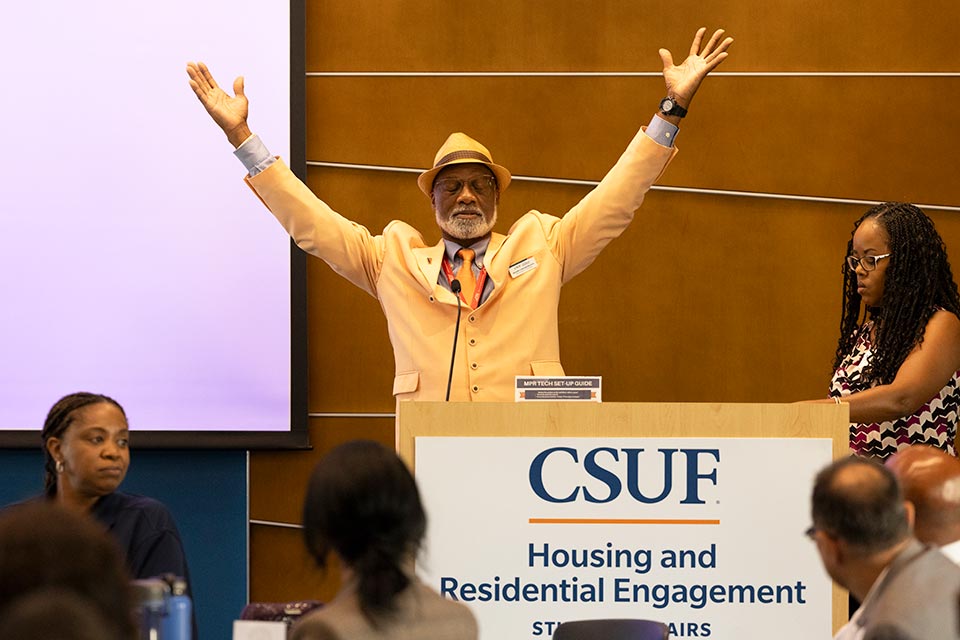 A speaker at a podium raises his hands in the air in dramatic gesture.